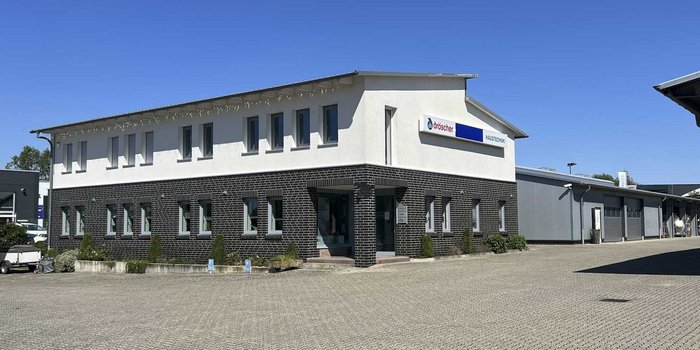 Ein modernes, zweigeschossiges Gebäude mit einer hellen Fassade und einem markanten schwarzen Sockel, umgeben von einem gepflasterten Parkplatz.